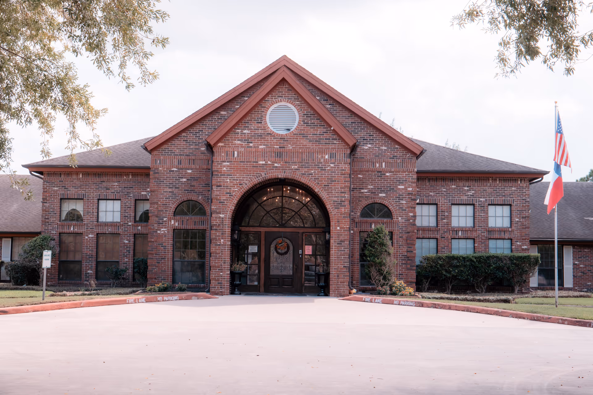 Front exterior view of a brick building with a large arched entrance door decorated with a wreath. The building has multiple windows and a triangular roof peak with a circular vent. There are two flags on the right side, one American and one Texas state flag. Shrubs and small trees are planted along the front of the building.
