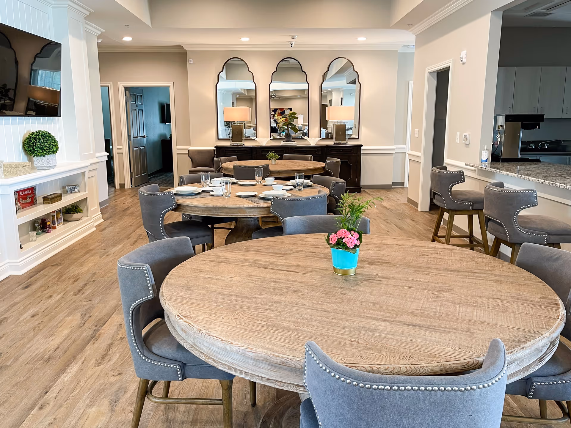 A bright and spacious dining area in a senior care facility featuring round wooden tables with gray upholstered chairs. The tables are set with plates, cups, and glasses. There are decorative plants on the tables and a sideboard with three large mirrors and lamps against the back wall. To the right, there is a kitchen counter with bar stools, and to the left, a wall-mounted TV above a white built-in shelf with board games and decorative items.