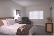 A clean, neatly made bedroom with a bed, chair, nightstand, and window blinds.
