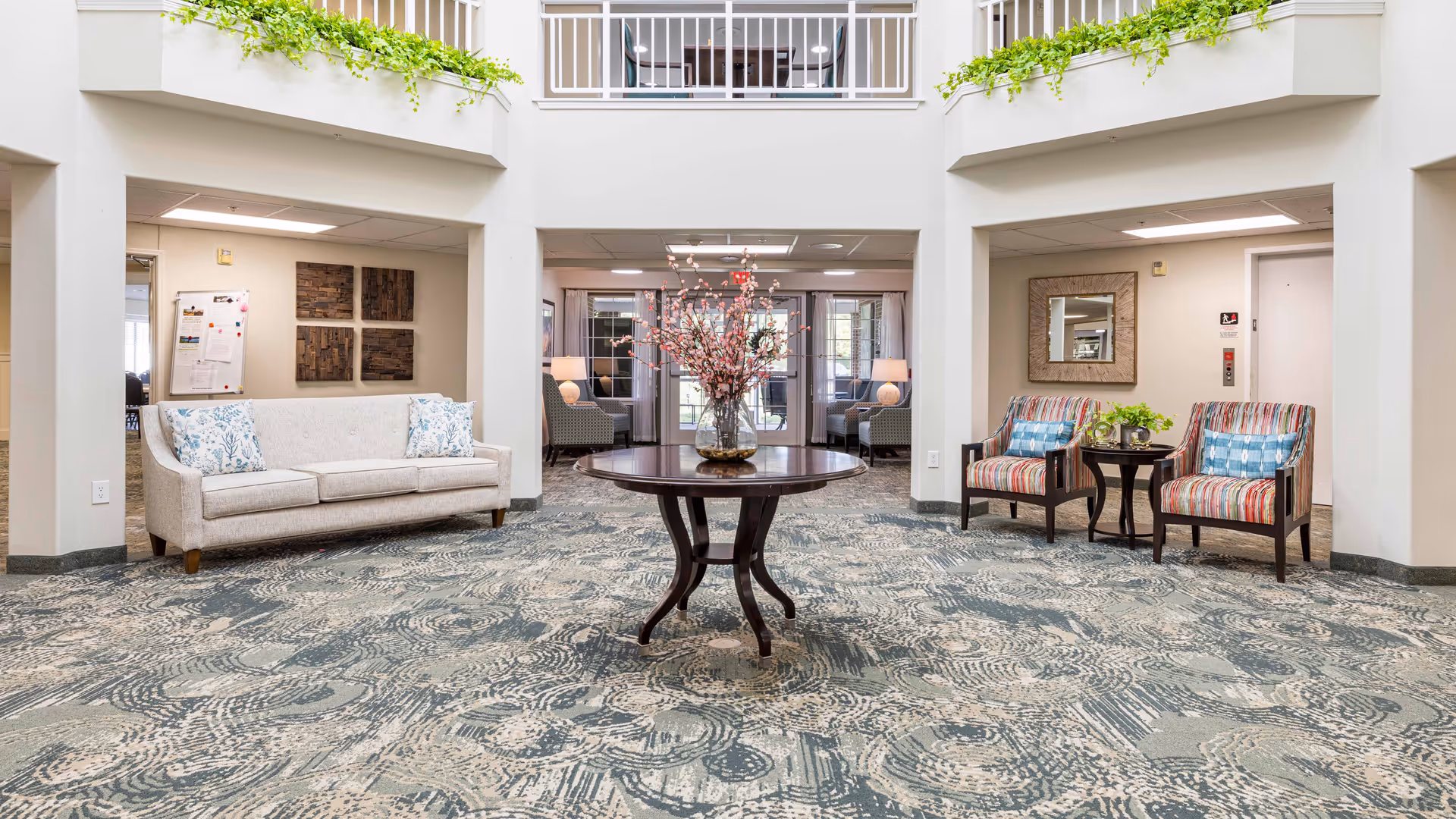 A bright and spacious common area in a senior living facility featuring a round wooden table with a floral arrangement in the center. The room has patterned carpet flooring, a beige sofa with floral cushions on the left, and two striped armchairs with blue pillows on the right, separated by a small round table with a plant. The area is surrounded by white walls and large windows, with a second-floor balcony and greenery above.