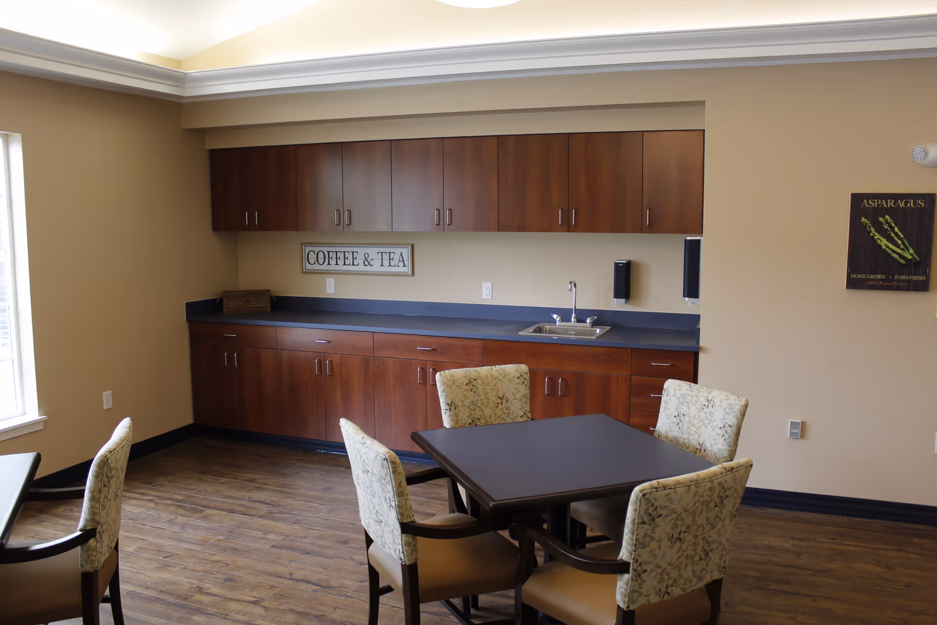 A small dining area with a square table surrounded by four cushioned chairs with floral upholstery. Behind the table is a kitchenette with dark wood cabinets, a countertop, a sink, and a sign on the wall that reads 'COFFEE & TEA'. The room has beige walls, a window on the left, and a wooden floor.