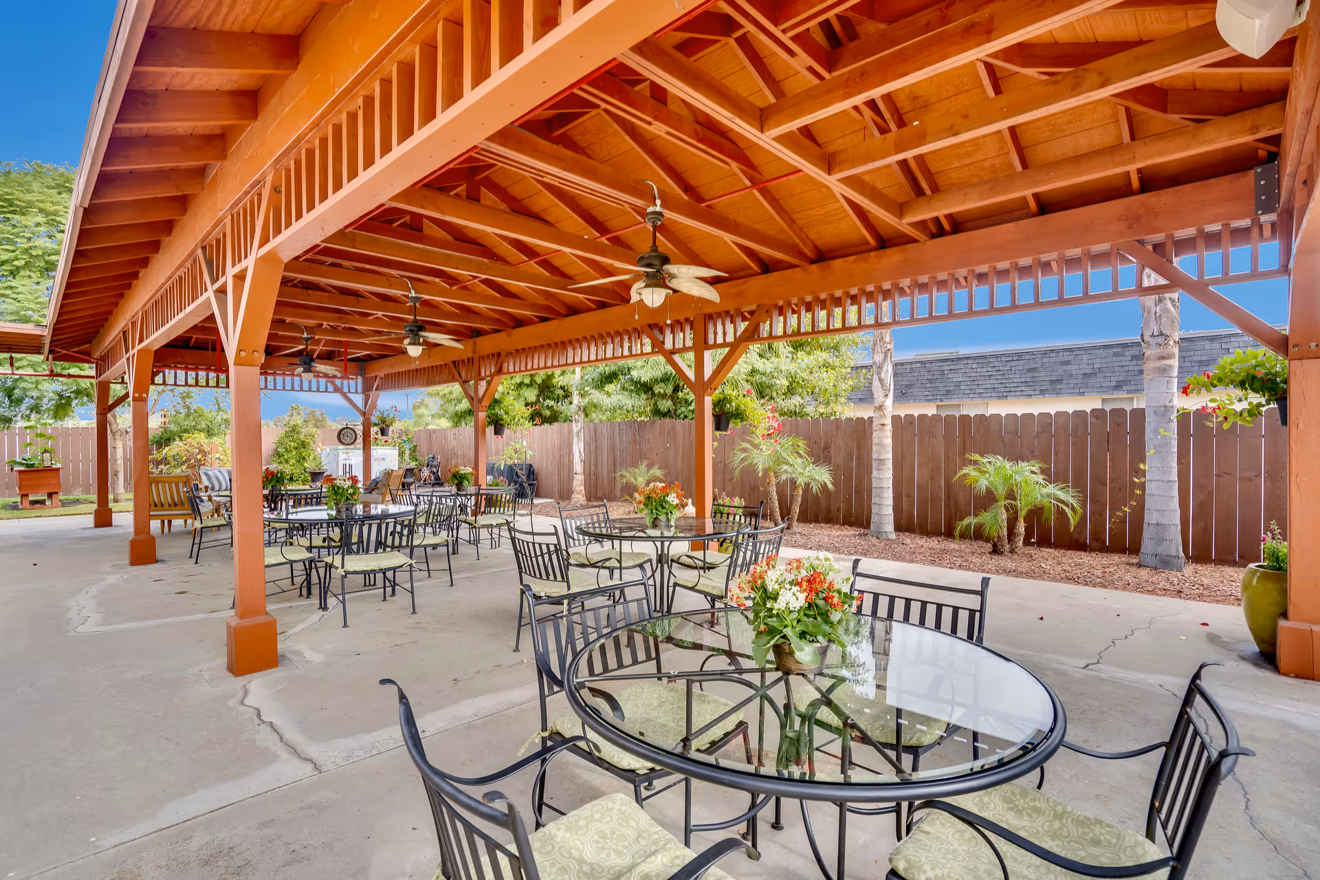 Outdoor covered patio area with multiple round glass tables and metal chairs with cushions. The patio has a wooden roof structure with ceiling fans. There are plants and flowers on the tables and around the patio, with a wooden fence and trees in the background under a clear blue sky.