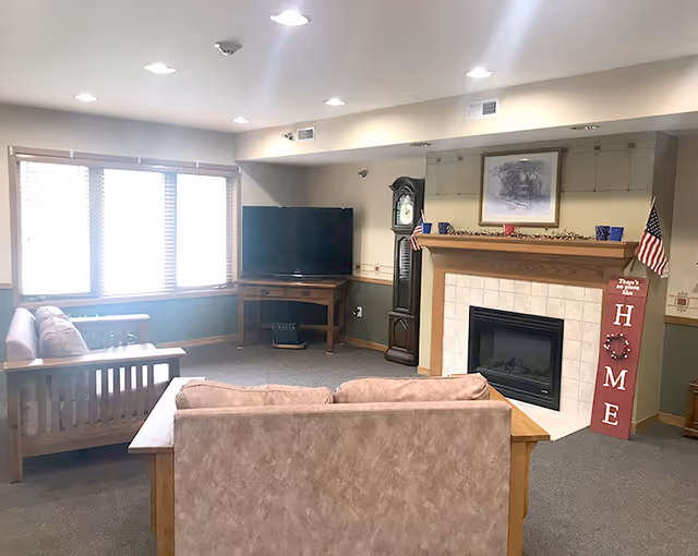 Cozy common room with sofas facing a fireplace and TV, a grandfather clock, and a large window letting in daylight.