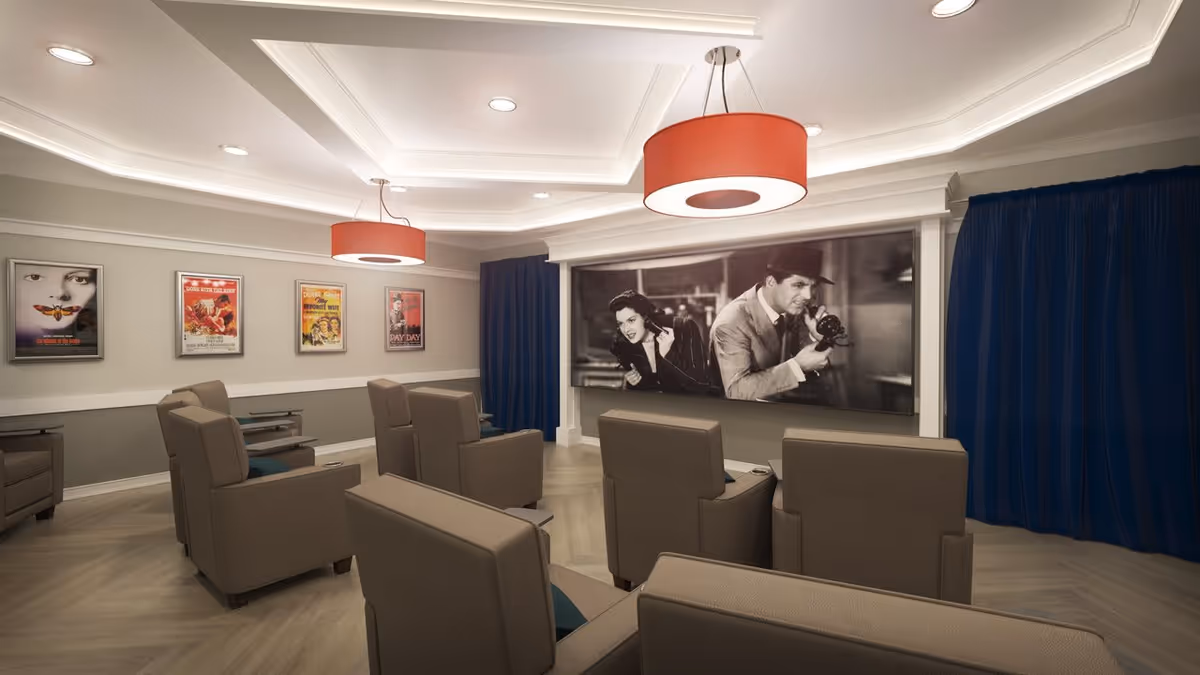 A small theater room with several brown recliner chairs arranged in rows facing a large screen showing a black-and-white movie scene. The room has light-colored walls with framed vintage movie posters and blue curtains on the sides. Two large red pendant lights hang from the ceiling.
