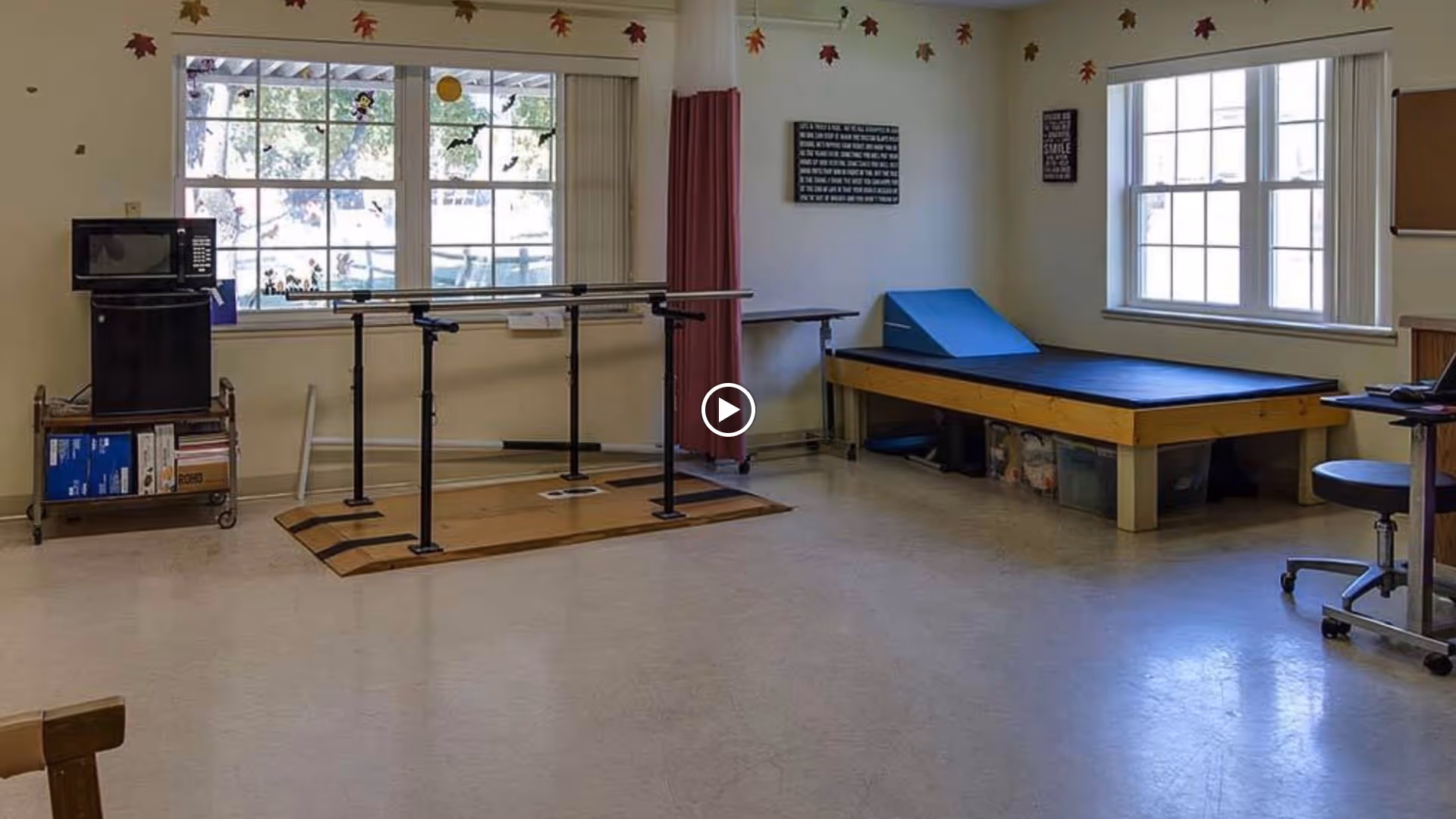 A therapy room with parallel bars for walking exercises, a padded therapy table with a blue wedge cushion, a microwave on a cart, and large windows letting in natural light. The room has a clean, spacious floor and motivational wall art.