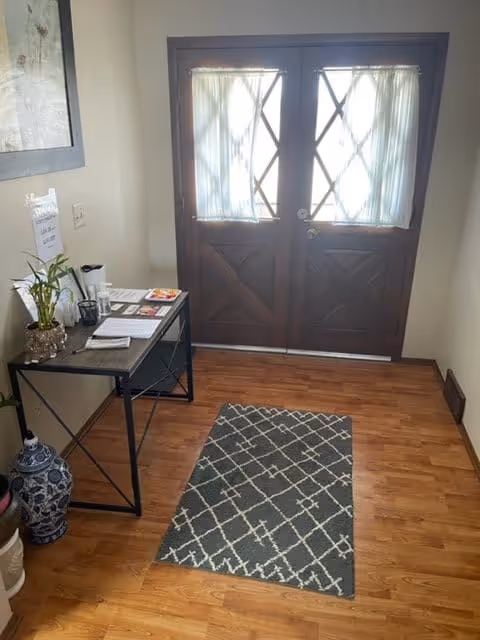 Entryway with double wooden doors featuring glass panels covered by sheer curtains. A small table on the left holds a plant, papers, and a cup. A patterned rug lies on the wooden floor in front of the doors. A large decorative vase is placed on the floor near the table.