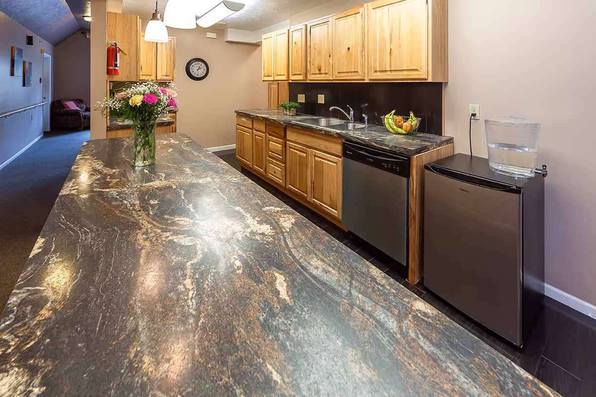 Communal kitchen area with long marbled countertops, wooden cabinets, sink, dishwasher, mini fridge and a vase of flowers.