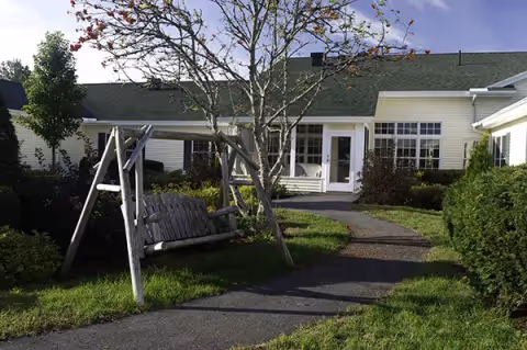 Outdoor courtyard showing a wooden swing bench, paved walkway, grassy landscaping and the entrance to a light-colored building.