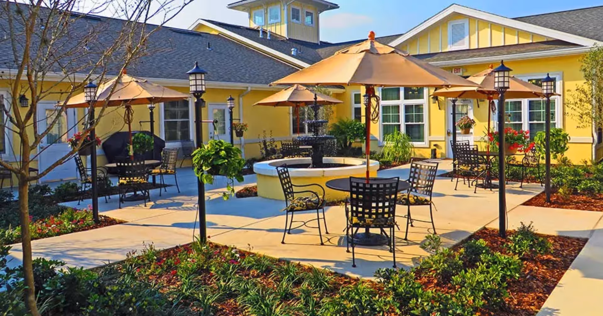 Outdoor patio area at Benton House of Clermont with several round tables and chairs under large beige umbrellas, surrounded by landscaped garden beds and lamp posts, adjacent to a yellow building with multiple windows.