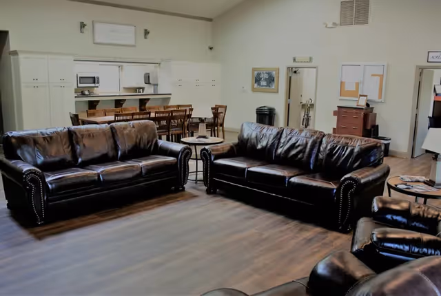 A spacious common area with two dark brown leather sofas and a matching armchair arranged around small round tables. In the background, there is a long wooden dining table with chairs, white cabinets, and a microwave. The room has light-colored walls and wood flooring.