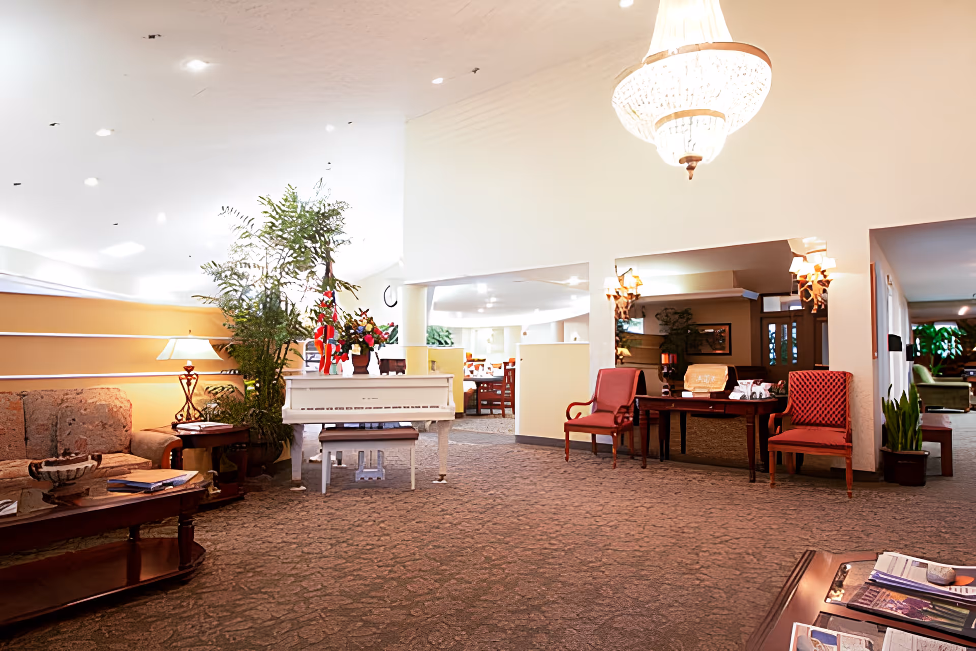 A spacious and well-lit living room area in a senior living facility featuring a white piano with floral arrangements on top, a patterned sofa, wooden coffee tables with magazines, red upholstered chairs, and various plants. The room has a carpeted floor, warm lighting from lamps and a chandelier, and an open layout leading to a dining area in the background.