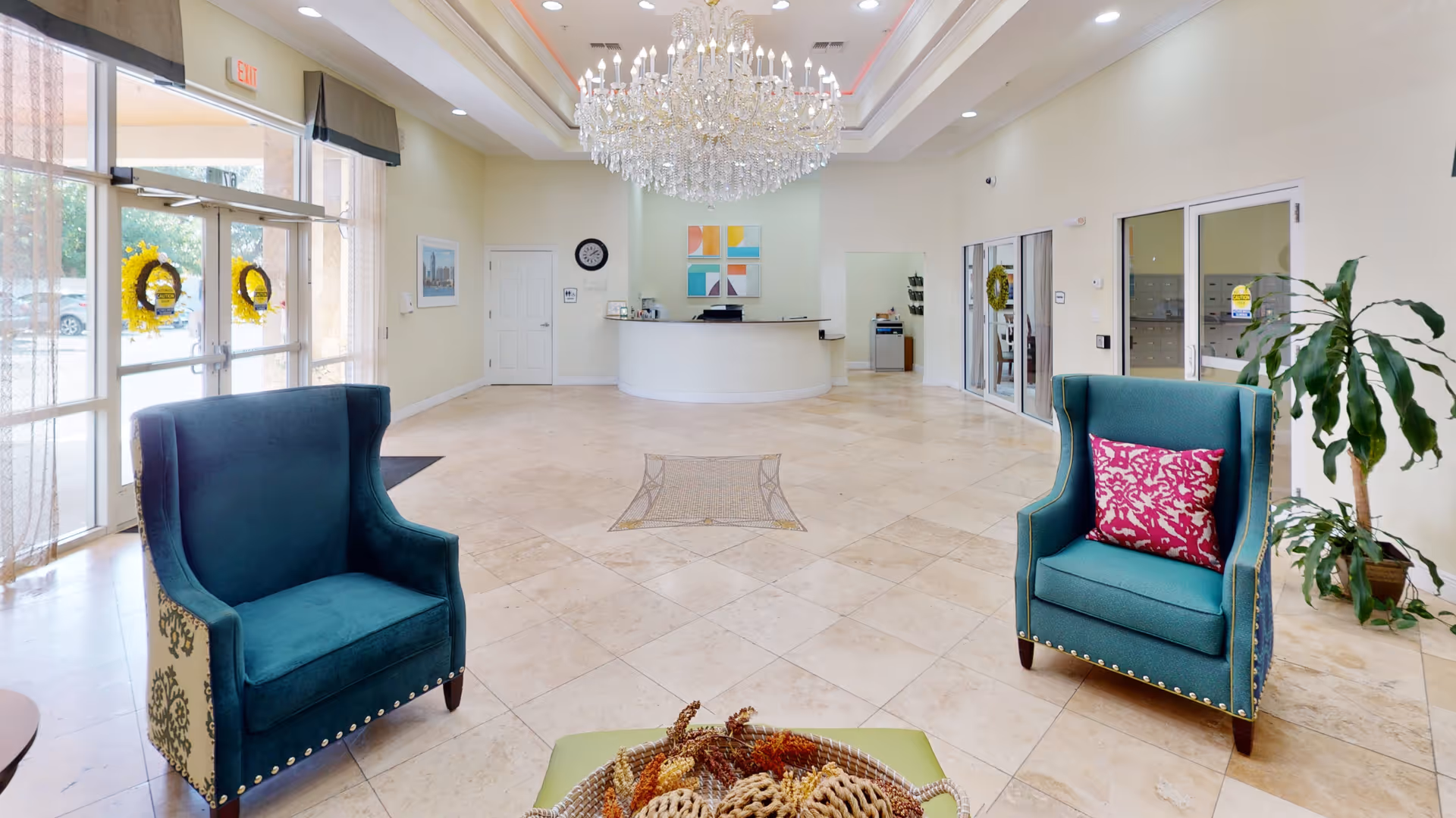 Spacious and well-lit lobby area with large glass entrance doors adorned with yellow wreaths. The room features a large chandelier hanging from the ceiling, a curved reception desk at the far end, two teal armchairs with decorative pillows, and a potted plant near the right wall. The floor is tiled with light-colored tiles, and there is a decorative inlay in the center of the floor.