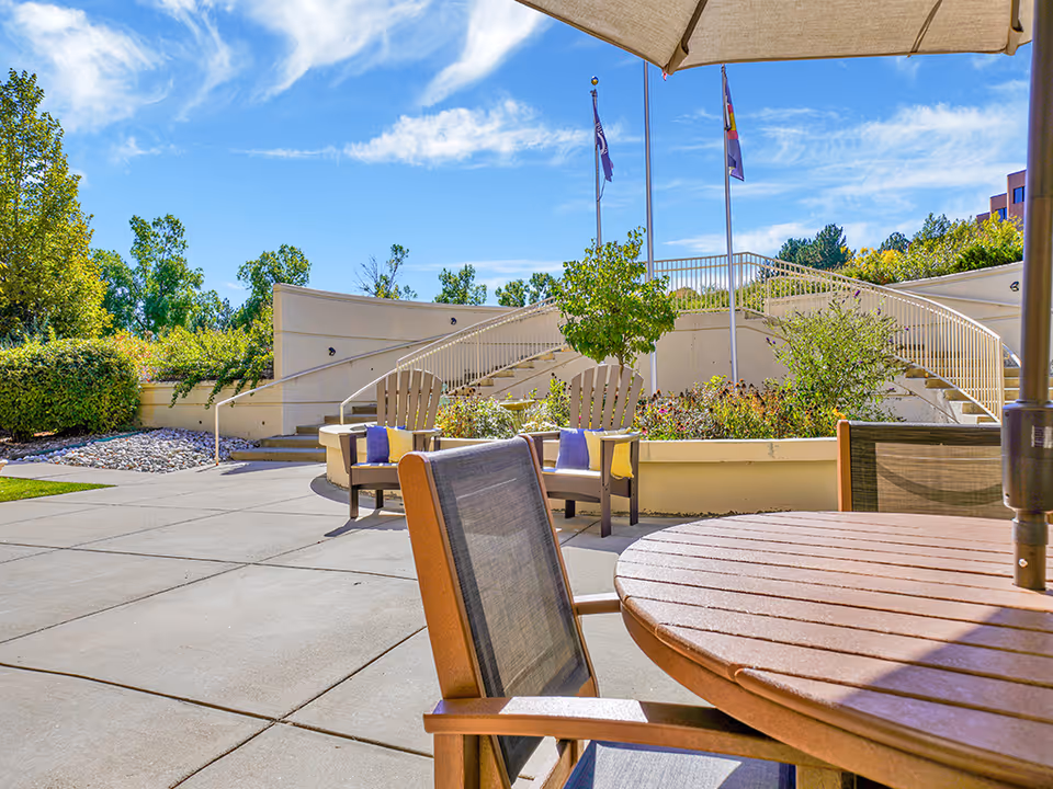 Outdoor patio with a round wooden table, chairs and umbrella overlooking landscaped steps and flagpoles under a blue sky.