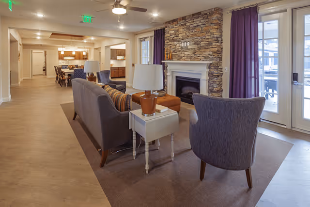 A cozy living room area with a stone fireplace, a gray sofa, two armchairs, a small white side table with a lamp, and purple curtains. The room opens into a dining area and kitchen in the background, with wooden cabinets and a dining table with chairs. Large glass doors lead to an outdoor space.