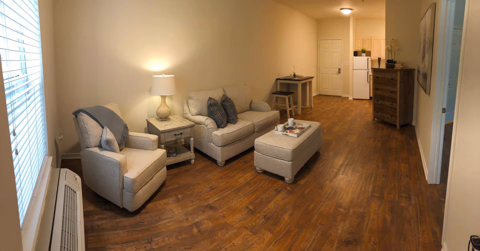 A cozy living area with a beige armchair and loveseat, a small wooden side table with a lamp, and an ottoman with a tray holding cups and a magazine. The room has wooden flooring and beige walls. In the background, there is a small kitchen area with a refrigerator, a chest of drawers, and a small dining table with two stools. A door is visible at the far end of the room.