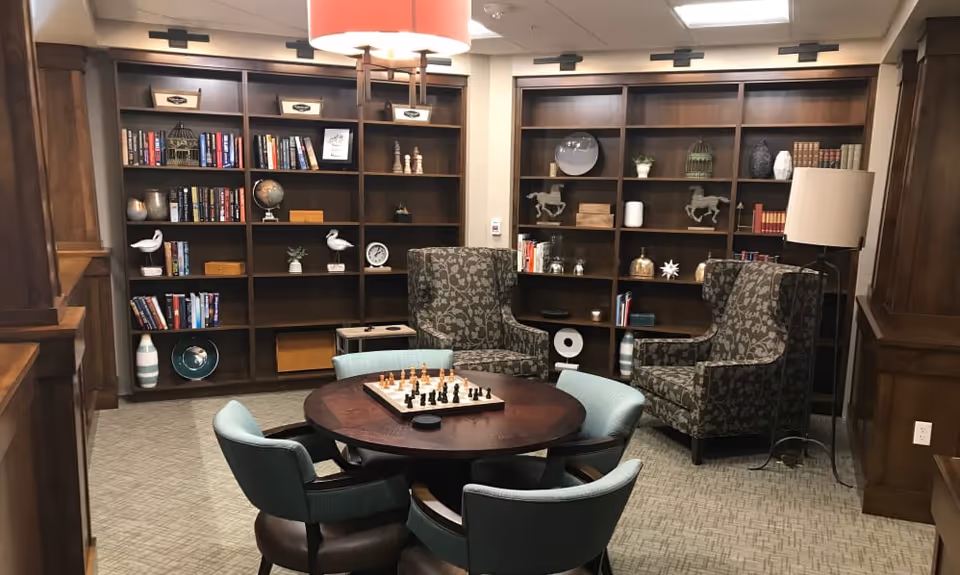 Cozy interior lounge with a round table and a chessboard, upholstered chairs and two wingback chairs arranged in front of built-in bookshelves.