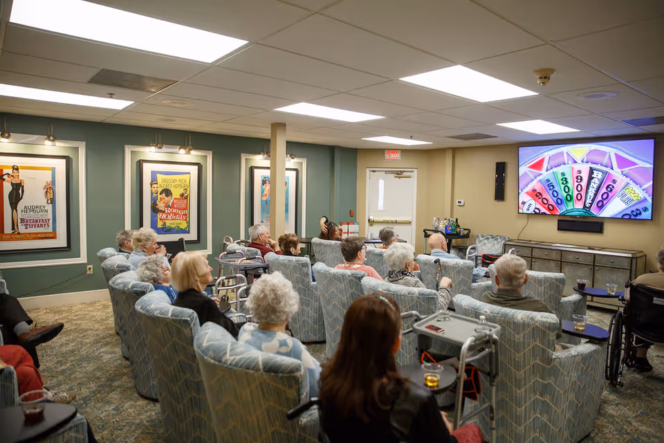 A group of elderly residents seated in upholstered chairs watching a large wall-mounted TV in a common activity room with framed posters on the walls.