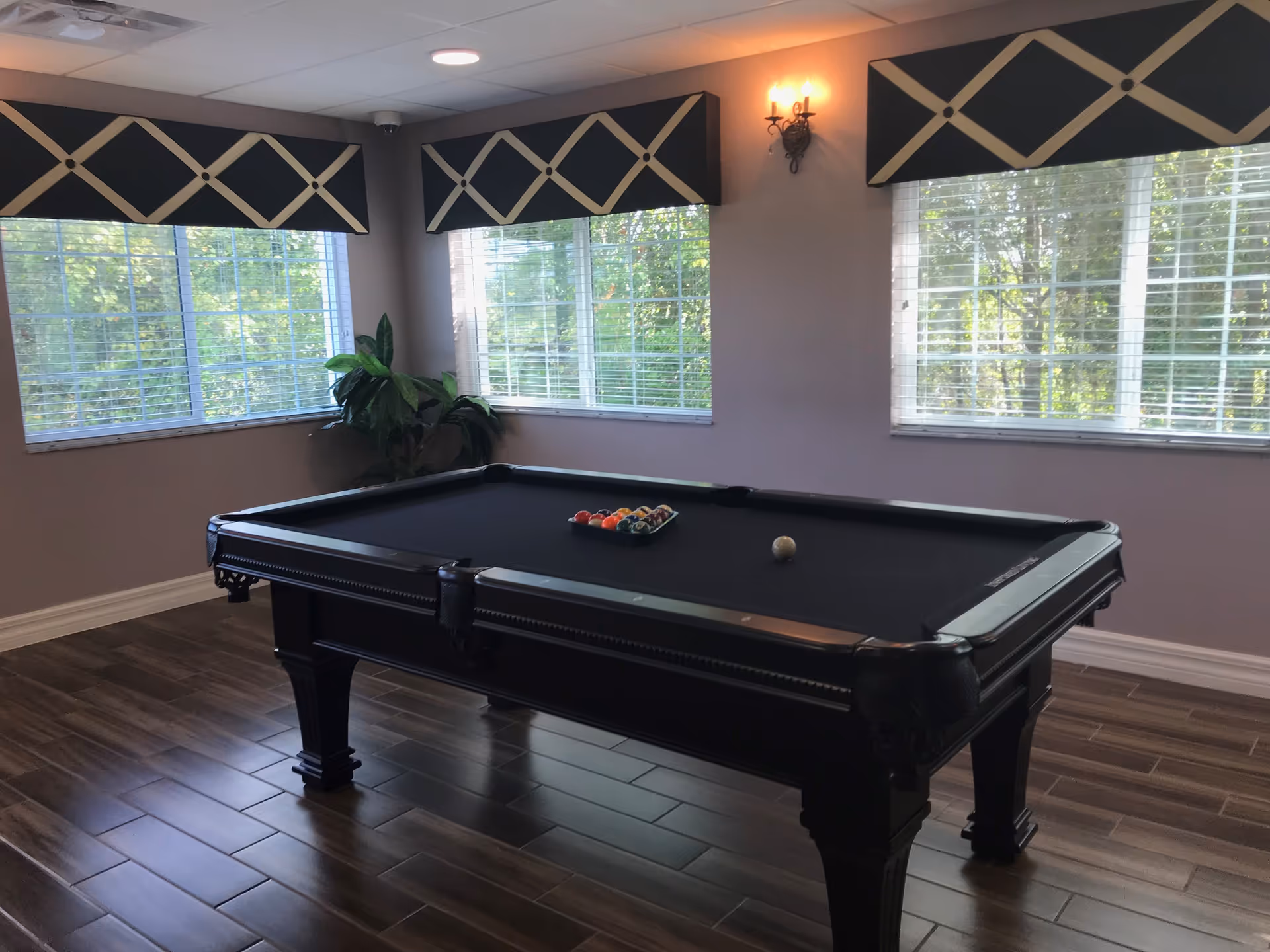 A room with a black pool table in the center, set up with billiard balls and a cue ball. The room has large windows with white blinds and decorative black and beige valances. There is a plant in the corner and a wall sconce light fixture on the wall. The floor is covered with brown wood-like tiles.