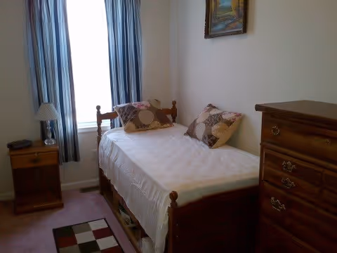 A small bedroom with a single wooden bed covered with a white bedspread and two patterned pillows. Next to the bed is a wooden nightstand with a lamp and a small item on it. There is a window with striped curtains letting in natural light. On the right side of the room is a wooden chest of drawers, and a framed picture hangs on the wall above the bed.