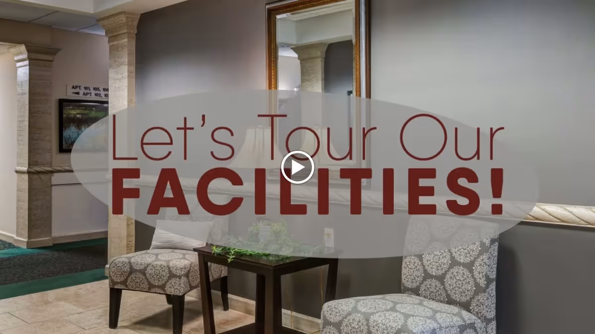 A seating area in a senior living facility with two patterned chairs and a small dark wooden table between them, a large mirror on the wall, and a hallway with stone columns and directional signs in the background.