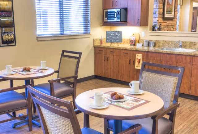 A cozy dining area with round tables set with white coffee cups and plates with pastries. The room features wooden chairs with cushioned seats, a countertop with a sink, cabinets, a microwave, and a small chalkboard sign displaying the coffee of the day. A window with blinds allows natural light to enter the space.