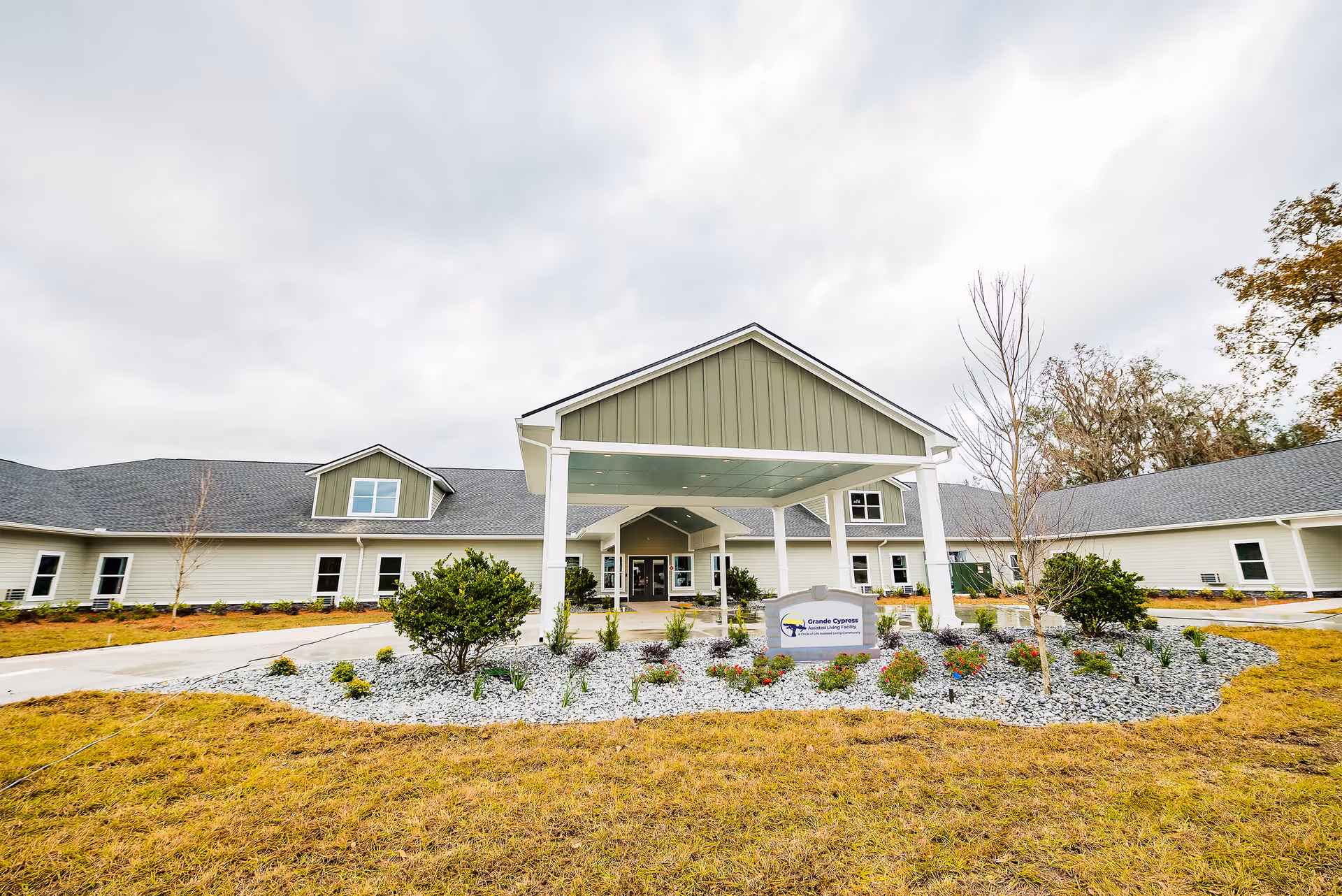 Front exterior view of Grande Cypress Assisted Living Facility showing a large covered entrance with a driveway, landscaped garden with small bushes and flowers, and a cloudy sky overhead.