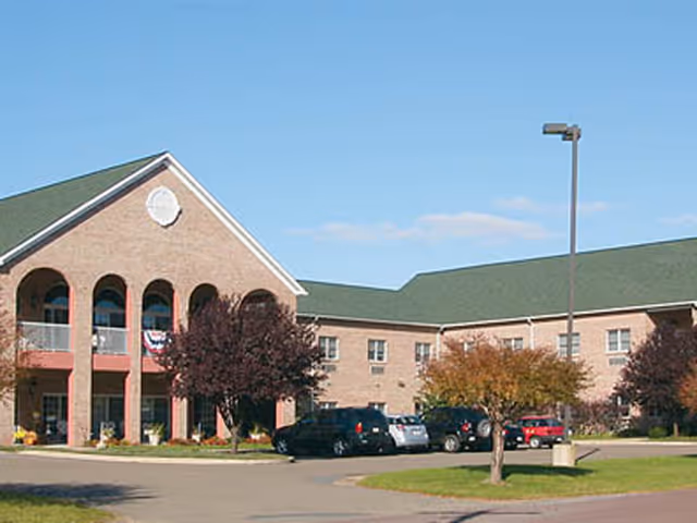 Exterior view of a two-story brick building with a green roof, arched windows, and a covered entrance. Several cars are parked in front of the building, and there are trees and a streetlamp in the parking area under a clear blue sky.