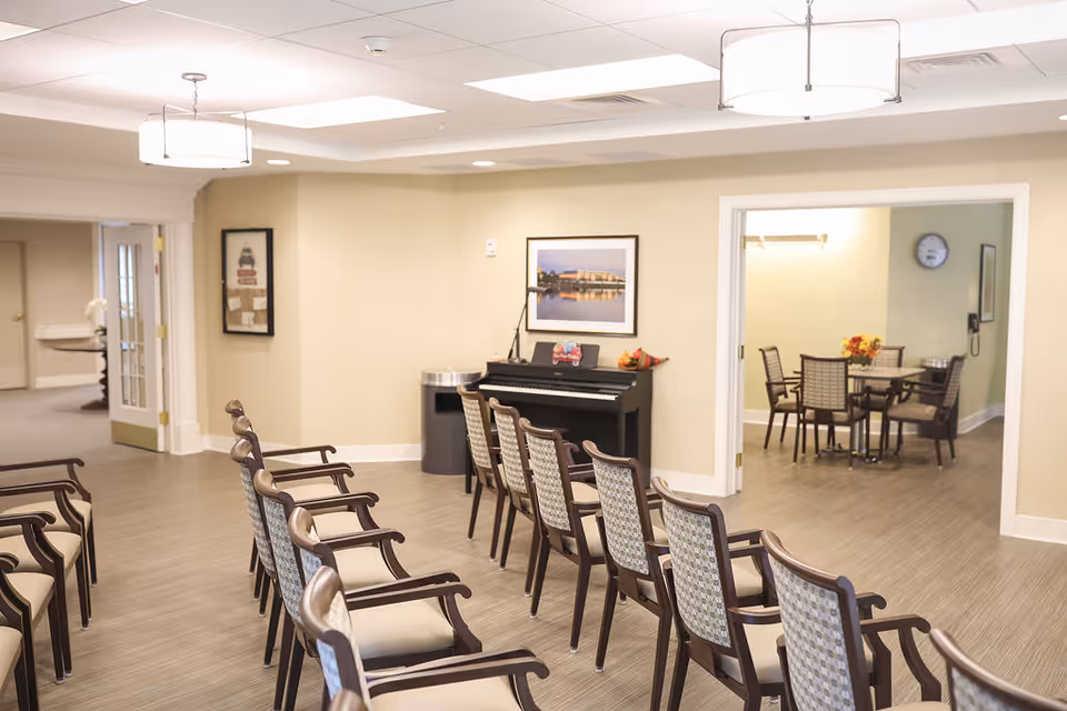A senior living facility common area with two rows of wooden chairs facing a black upright piano against a beige wall. A framed picture hangs above the piano, and a trash can is nearby. Through an open doorway, a smaller room with a round table and four chairs is visible, decorated with a vase of flowers and a wall clock.