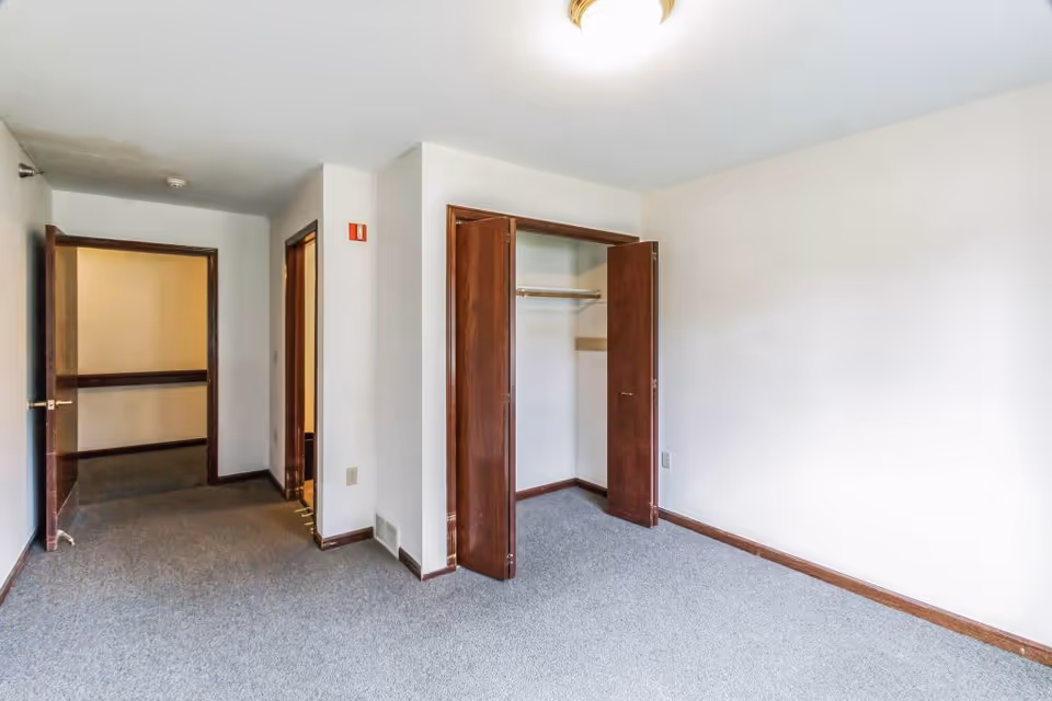 Empty room with light gray carpet, white walls, an open wooden closet with double doors, and an open doorway leading to a hallway.