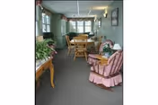 A cozy interior room with green walls featuring a wooden rocking chair with a pink cushion and skirt, a small table with a lamp and flowers, a wooden dining table with chairs, and large windows with curtains letting in natural light.