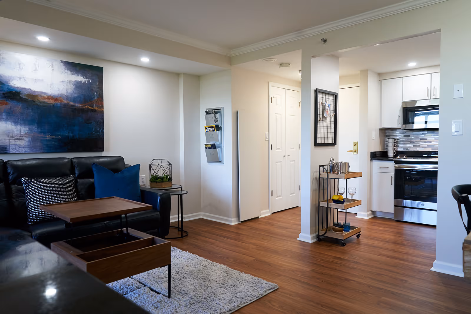 A modern living area with a black leather sofa adorned with blue and patterned pillows, a wooden coffee table with a lift-top, a small round side table with a decorative plant, and a large abstract painting on the wall. The room has wood flooring and leads into a kitchen area with white cabinets, stainless steel appliances, and a small rolling cart with glassware and a pitcher.
