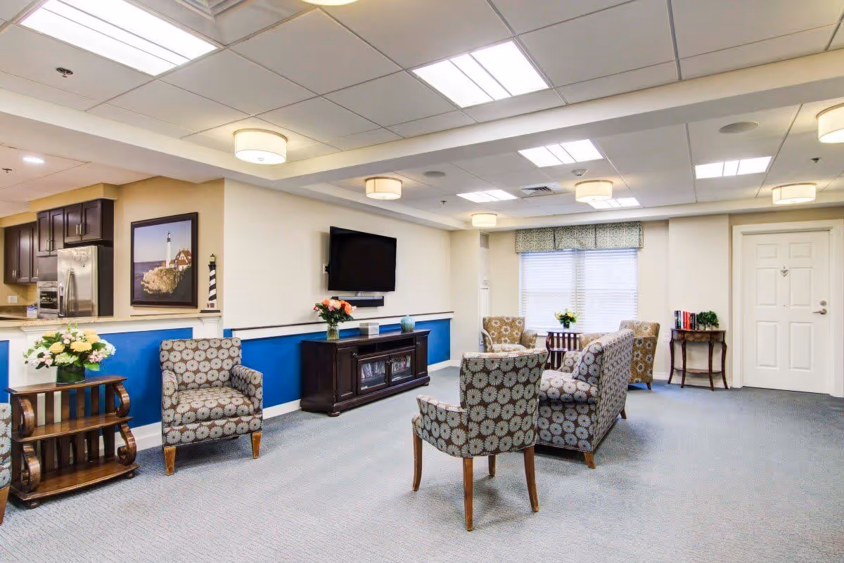 A bright and spacious living room area in a senior living facility with patterned armchairs and a sofa arranged around a dark wooden TV stand with a flat-screen TV mounted on the wall. The room features a blue accent wall, a window with a valance, ceiling lights, and a small table with flowers and books near the entrance door. The kitchen area with stainless steel appliances is partially visible on the left.