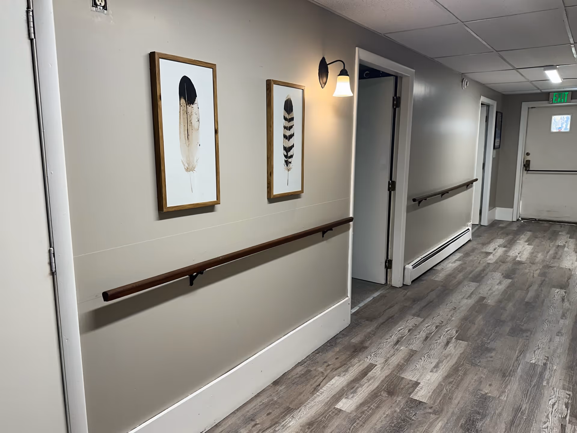 A clean hallway in a senior living facility with wood-like flooring, beige walls, two framed feather artworks, wooden handrails on both sides, a wall-mounted light fixture, and several doorways leading to other rooms. An exit door is visible at the end of the hallway.