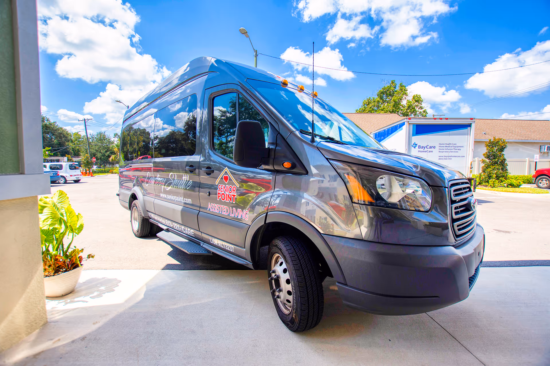 A gray Senior Point Assisted Living van parked outside on a sunny day with a blue sky and scattered clouds. The van has the Senior Point Assisted Living logo and contact information on its side. There are some plants in a pot near the entrance and other vehicles and buildings in the background.