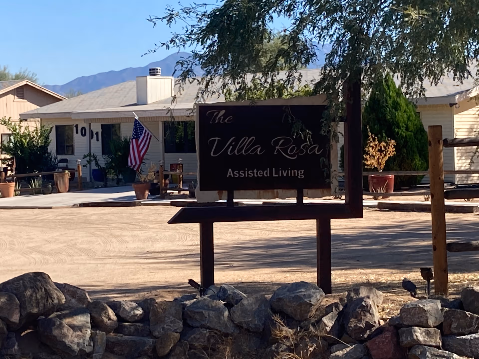 Wooden sign reading "The Villa Rosa Assisted Living" in front of a single-story building with an American flag and desert landscaping.