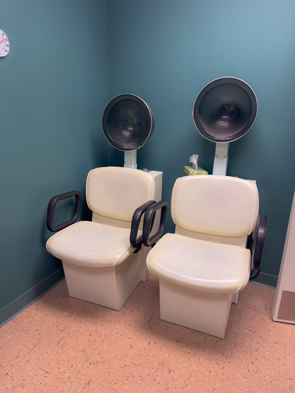 Two vintage salon hair dryer chairs with cream-colored seats and black armrests against a teal wall in a small room with a clock on the wall.