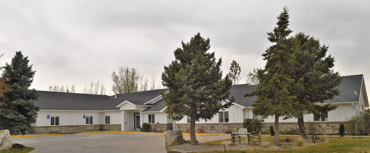 Exterior view of Oak Ridge Assisted Living facility showing a single-story building with white siding and a dark roof. The entrance is centered with a small covered porch. There are several large evergreen trees and some landscaping with rocks and grass in front of the building. The sky is overcast.