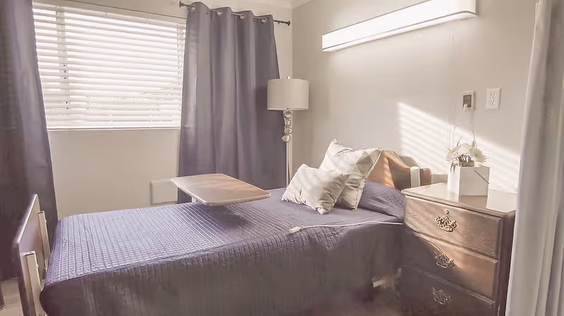 A small bedroom with a single bed covered in a dark quilted bedspread, two pillows, and a wooden overbed table. Next to the bed is a wooden nightstand with a small flower arrangement and a lamp with a tall stand. The room has a window with white blinds and dark curtains.