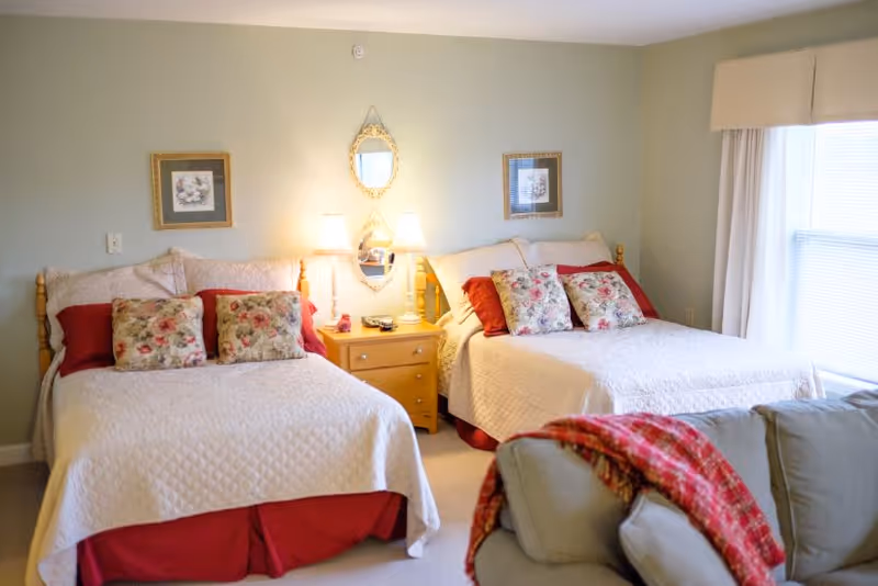 A cozy bedroom with two double beds, each adorned with white quilts, red bed skirts, and floral pillows. Between the beds is a wooden nightstand with two lit lamps and decorative items. Above the nightstand hangs a small ornate mirror flanked by two framed pictures. A window with blinds and a beige valance is on the right side, and a gray couch with a red plaid blanket is partially visible in the foreground.