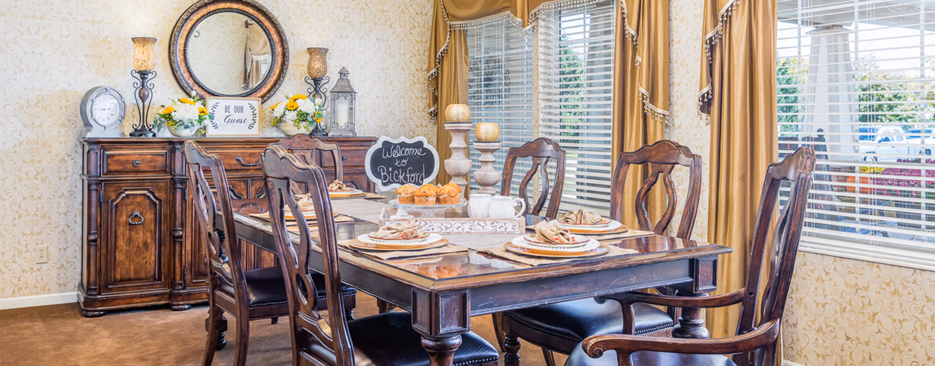 A warmly decorated dining room with a wooden dining table set for four, featuring plates, napkins, and a centerpiece with muffins and candles. Behind the table is a wooden sideboard with decorative items including a clock, flowers, lamps, and a round mirror. A small chalkboard sign on the table reads 'Welcome to Bickford'. Large windows with gold curtains allow natural light into the room.