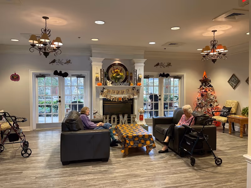 A senior living facility lounge with a fireplace, seasonal decorations, and two residents seated on sofas.