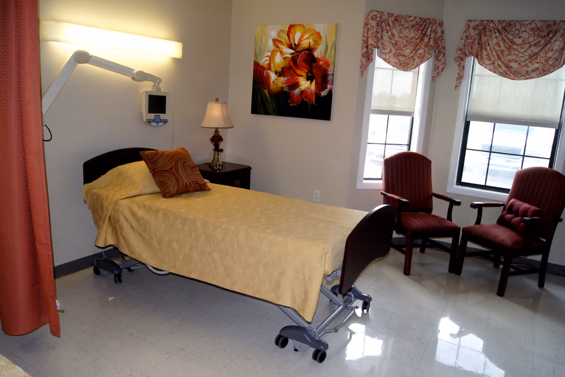 A nursing facility room with a single hospital bed covered in a yellow bedspread and a decorative pillow. There is a bedside table with a lamp, a wall-mounted medical device, two red upholstered chairs near two windows with patterned valances, and a colorful floral painting on the wall.