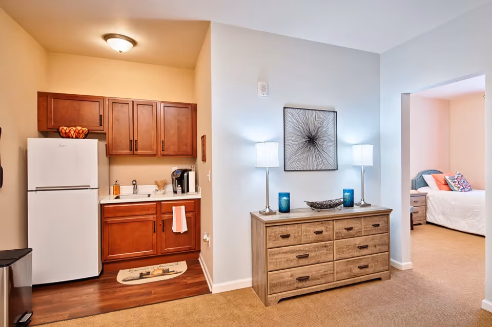 Small apartment interior with a kitchenette on the left, a wooden dresser with lamps in the center, and a bedroom visible through an open doorway.