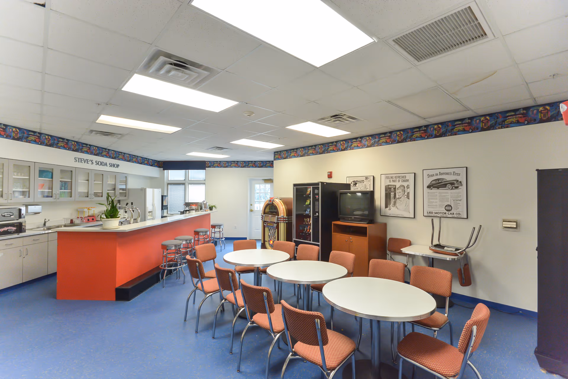A bright and clean common room with a retro soda shop theme named 'Steve's Soda Shop'. The room features a long counter with stools, several round tables with orange cushioned chairs, a jukebox, a vending machine, and a TV on a wooden stand. The walls are decorated with vintage-style posters and a colorful border near the ceiling.