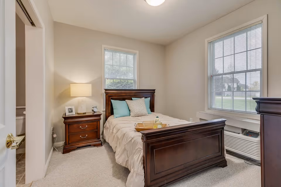 Cozy bedroom featuring a dark wood bed and nightstand with a lamp, two windows, and light-colored walls.