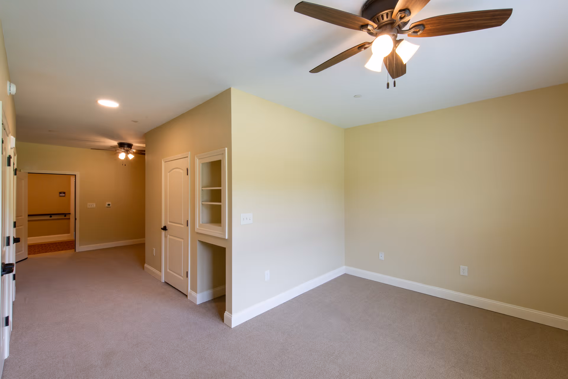 An empty room with beige walls and carpeted floor, featuring a ceiling fan with lights. There is a small built-in shelving unit and a closed door on one wall. The room opens into a hallway with another ceiling fan and light visible in the distance.
