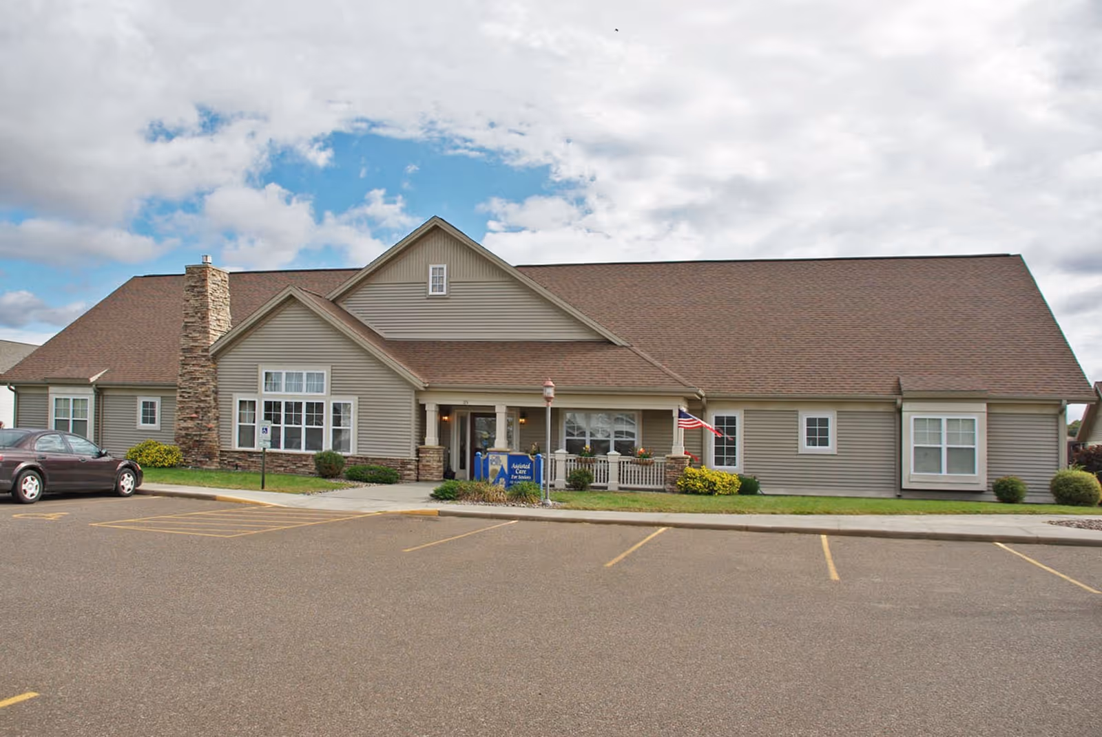 Exterior view of a single-story senior living facility building with beige siding and a brown roof. There is a stone chimney on the left side, a small porch with white railing near the center, an American flag displayed, and a parking lot in front with marked spaces. The sky is partly cloudy with patches of blue.