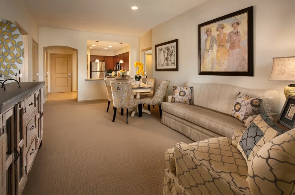 Open living and dining area with a sofa, patterned armchair, round dining table, and a kitchen visible in the background.