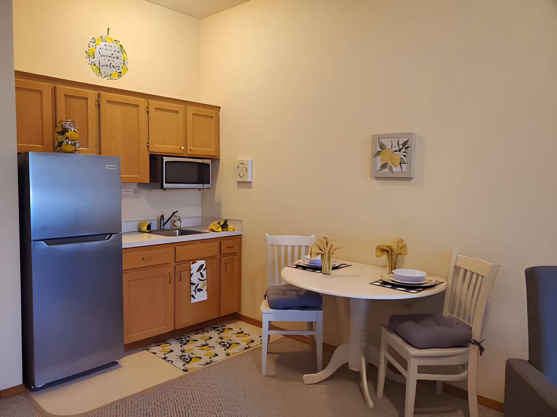 Small kitchen area with wooden cabinets, a stainless steel refrigerator, a microwave, and a sink. Next to the kitchen is a small round dining table set for two with white chairs and yellow-themed decorations including napkins and placemats. The walls are light-colored with lemon-themed artwork.