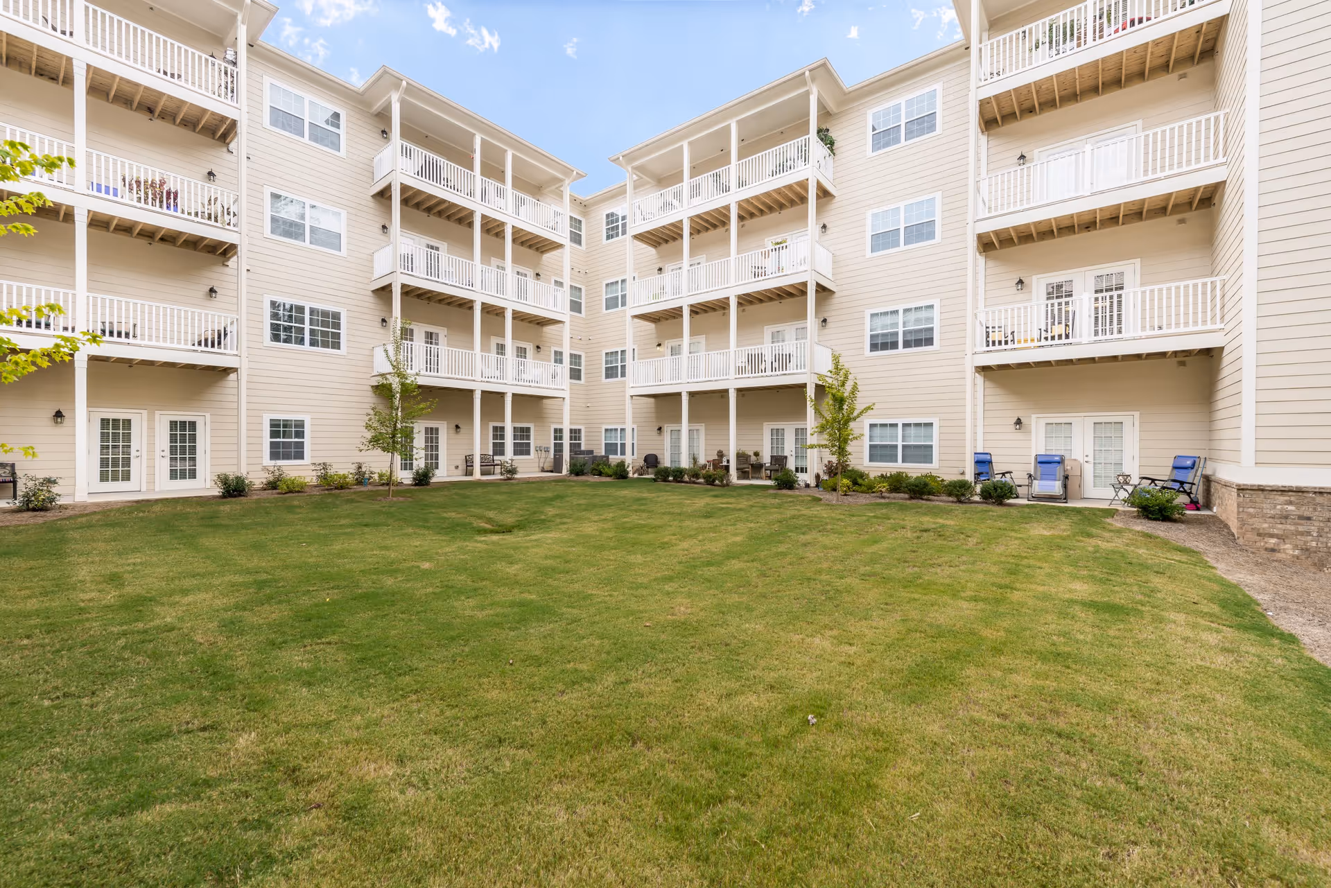 View of a senior living facility courtyard with a well-maintained green lawn surrounded by a beige multi-story building featuring balconies and patio doors. Some outdoor chairs and small tables are placed near the ground floor entrances.